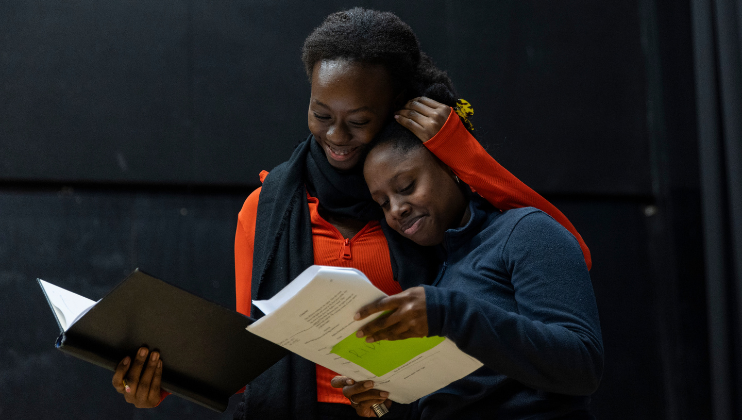 Two individuals stand close together, smiling warmly as they read from scripts. The person on the left, wearing an orange jacket and black scarf, embraces the other, who is dressed in a dark sweater and holding a green script. They appear to be in a theater setting.