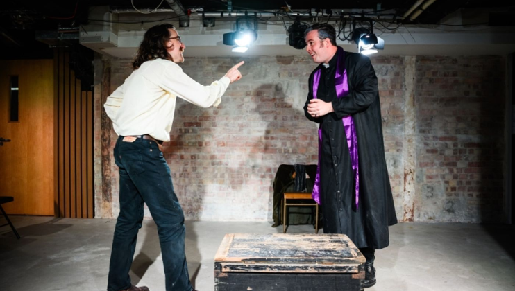 James Demaine, in character as Daniel Farringdon, stands one side of a suitcase and wags his finger at Alex Knott, the other side of the suitcase and in character as Bishop Patrick Sheffield