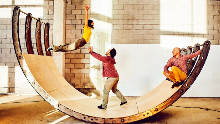 A semi-circular structure, like the surface of a halfpipe without a foundation, leans to one side in a gym; one the leaning side, a man sits watching as a child jumps from the other side into the arms of a man standing in the middle of the structure