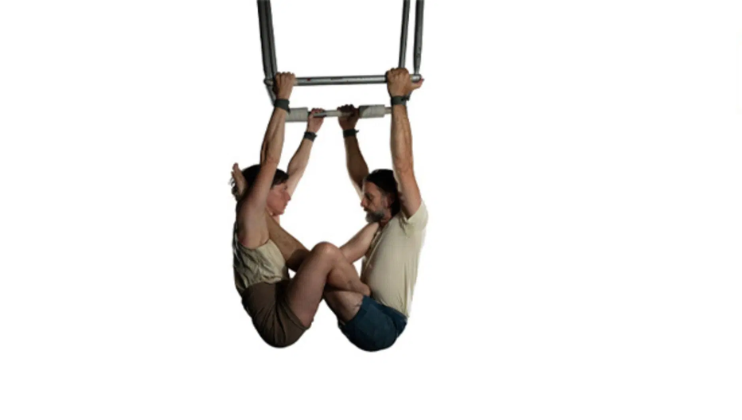 Against a blank white background: Charlotte Mooney and Alex Harvey, co-Artistic Directors of circus company Ockham's Razor, hang by their arms from a sparse metal frame facing one another. Each of them has their feet hoisted above the other's shoulders in in intimate position.