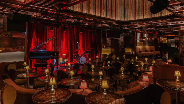 A view of the empty stage at Upstairs at Ronnie's from a few rows back; each circular table in descending rows towards the stage is gorgeously illuminated by a table lamp