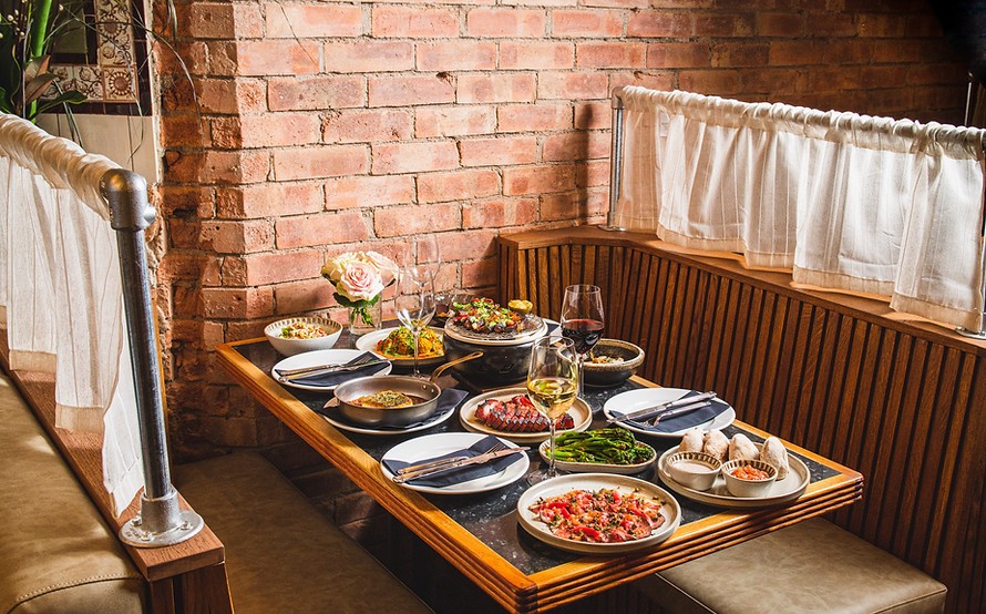 A cozy restaurant booth features a table laden with a variety of gourmet dishes. The assortment includes colorful salads, pasta, seafood, and side dishes. Two glasses of wine, one red and one white, are on the table. Brick walls and soft lighting add to the warm ambiance.