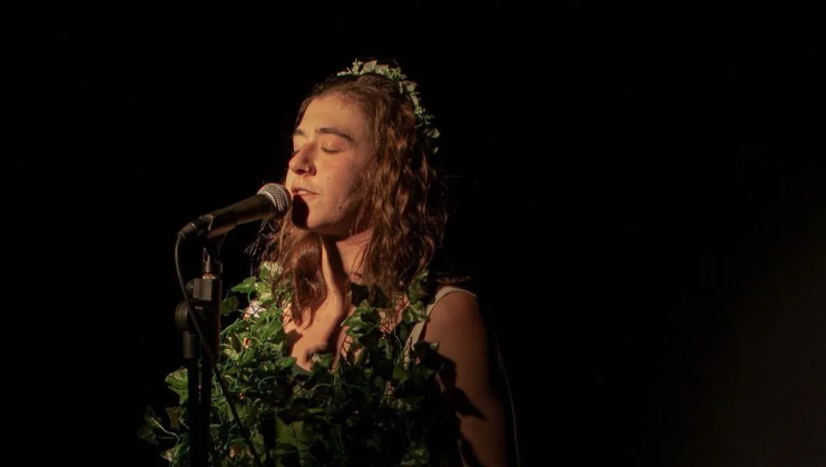 A close-up on Sammy Trotman in a green headdress. Eyes closed, she stands inches away from a microphone against a black background