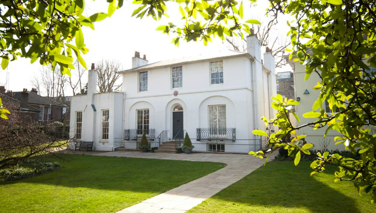 A two-story white house with a flat roof, large windows, and a central entrance, surrounded by greenery. A stone pathway leads to the front door. The house has a well-maintained lawn and is framed by leafy branches in the foreground.