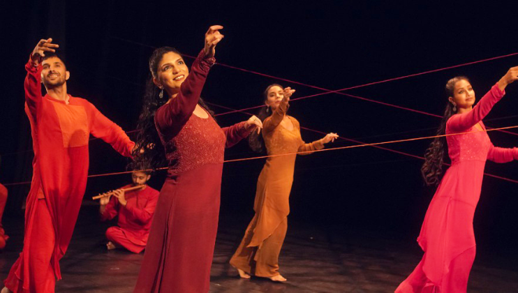 Four Kathak dancers - one male and three female from left to right - pose in traditional red and orange dress with one hand extended upwards