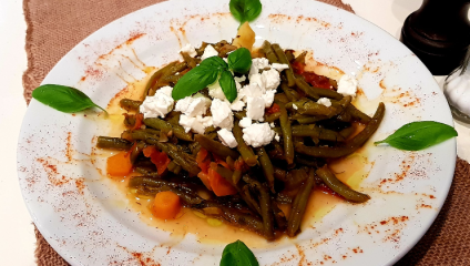 A white plate filled with a cooked green bean and tomato dish, topped with crumbled white cheese, garnished with fresh basil leaves. The plate sits on a brown woven placemat, and a light dusting of seasoning is visible around the edges of the dish.