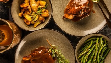 A top-down view of a meal with three plates. One plate contains roasted potatoes garnished with herbs, another has a meat dish with sauce and herbs, and the third plate has cooked green beans. A drink with a lemon slice is visible on the side.