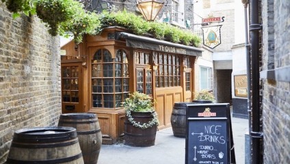 A charming, narrow alleyway leads to a traditional English pub with a wooden facade and large windows. The sign above reads Ye Olde Mitre. Barrels and potted plants decorate the entrance, and a chalkboard sign advertises drinks and beers.