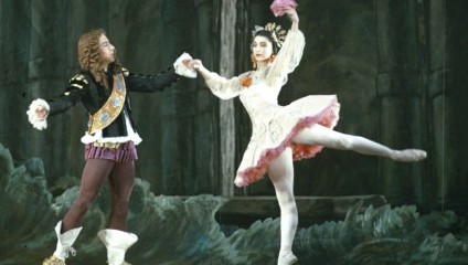Two ballet dancers perform on stage. One dancer, dressed as a prince, holds the hand of another dancer, who is poised on one leg in an elegant stance while wearing a white tutu and a tiara. The background features a dark, forest-like stage set.