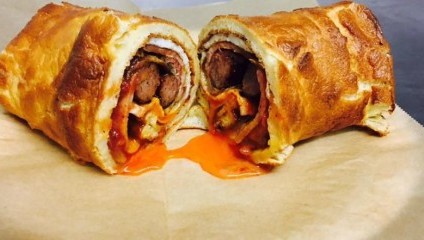 A close-up of a sliced sausage roll on parchment paper. The sausage roll has a golden-brown, flaky pastry exterior, and the filling includes sausage and a generous amount of vibrant red sauce, oozing slightly from the center.