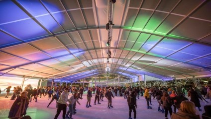 A large crowd of people is ice skating inside a spacious, covered rink. The roof is illuminated with colorful lights, creating a vibrant atmosphere. The rink is bustling with skaters of all ages, enjoying the lively, festive environment.