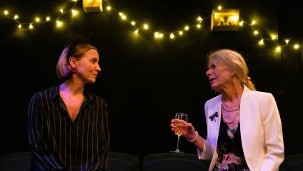 Two women are sitting and conversing. The woman on the left wears a dark, striped top with a headband. The older woman on the right is dressed in a white blazer over a floral dress, holding a wine glass. They appear to be on a stage set with string lights in the background.