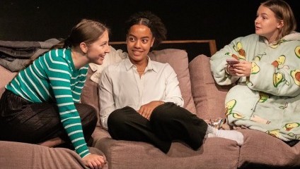 Three individuals are seated on a brown couch. The person on the left, wearing a green striped sweater, is talking and smiling towards the person in the middle, who is wearing a white shirt and has an arm resting on the back of the couch. The person on the right, in an avocado-print blanket, looks on. Above them are four white stars and the text London Theatre.