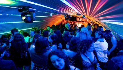 A crowded nightclub with vibrant, multicolored lights radiating from the ceiling. People are dancing, mingling, and enjoying the energetic atmosphere. The room is packed with individuals, some holding drinks, and a DJ booth is visible in the background.