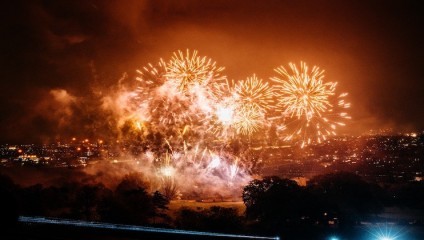 A vibrant display of fireworks illuminates the night sky over a cityscape. Multiple bursts of bright colors light up the scene, contrasting against the dark, smoky background. In the foreground, silhouettes of trees and buildings are visible.