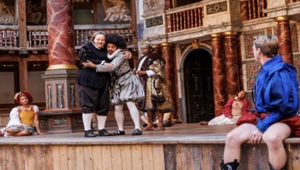 Actors perform a Shakespearean play on a traditional stage. Two characters in historical costumes embrace, while three others watch. One sits on the floor and another person, wearing a red and blue outfit, sits on the right edge of the stage.