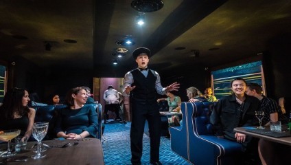 A man wearing a cap and vest passionately performs in the center of a dimly lit room, holding the attention of seated audience members at tables adorned with drinks. The room has a cozy, intimate atmosphere with people watching attentively.