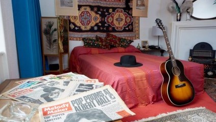A vibrant bedroom with a red bedspread, a black hat on the bed, and a decorated wall tapestry. Newspapers, including one with the headline Pop Proms at Albert Hall, are spread on a table. A guitar rests against the bed, and native headdresses adorn the walls.