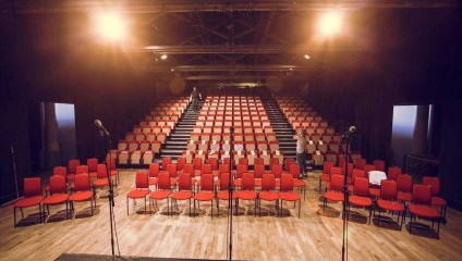 A view of an empty auditorium with red chairs arranged in rows, facing a stage with two microphones on stands. Bright stage lights illuminate the scene from the ceiling, highlighting the wooden floor and the symmetrical seating arrangement.