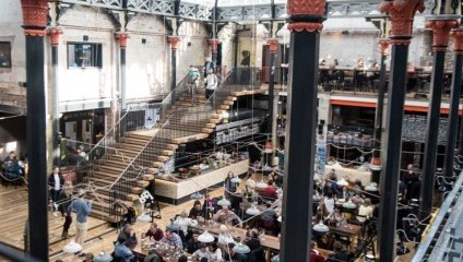 large Victorian market space with glass ceiling. Lots of people sitting at ground level on tables and chairs drinking and eating.