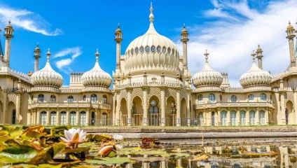 A grand, white palace with ornate domes and minarets, reflecting Indo-Saracenic architecture, stands against a vibrant blue sky. A pond with lily pads and flowers lies in the foreground, mirroring parts of the intricate building.
