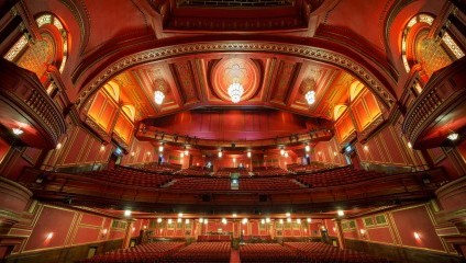 A grand, ornate theater with a red and gold color scheme, viewed from the stage. The design features a large, intricate ceiling with a central chandelier. Multiple balconies with red seats are visible, along with elegant architectural details and lighting fixtures.