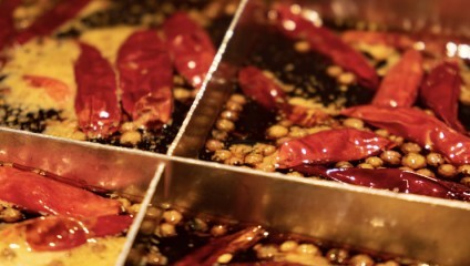 A close-up of a divided hot pot filled with a rich, dark broth. The hot pot sections contain red chili peppers and small round ingredients, likely peppercorns. The broth has a glossy, oily surface and appears very spicy.