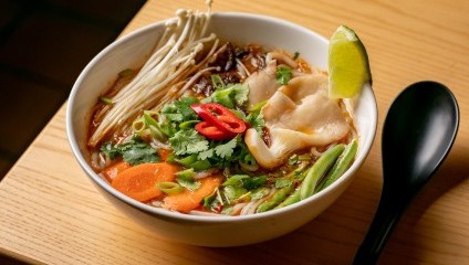 A bowl of ramen sits on a wooden table, filled with broth, noodles, enoki mushrooms, sliced pork, carrot slices, green onion, cilantro, and chopped red chili. A lime wedge garnishes the dish, and a black spoon rests beside the bowl.