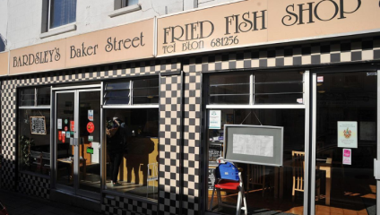 A storefront with signs reading Bardsley's Baker Street Fried Fish Shop. The exterior is decorated with a black and white checkered pattern. There are large glass windows, some with menus and notices displayed. A blue chair and standing sign are outside the entrance.