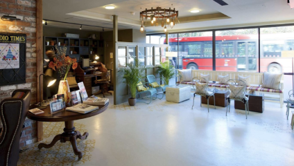 A cozy, well-lit café with a mix of vintage and modern decor. There are armchairs, a round wooden table with magazines, and a small shelf against a brick wall. Large windows show a passing red double-decker bus outside. The atmosphere is welcoming and relaxed.