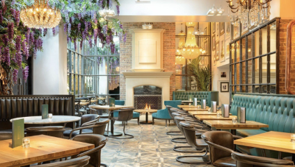 A stylish restaurant interior featuring wooden tables, black leather chairs, a large white fireplace, and a wall of windows. Crystal chandeliers hang from the ceiling, and vibrant purple flowers cascade from above, adding a touch of elegance to the space.