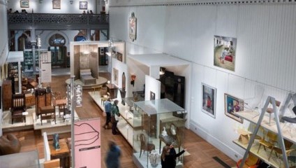A spacious art gallery with high ceilings, displaying various paintings, sculptures, and furniture. Visitors walk through the room observing the exhibits. A second-floor balcony provides additional display space and an overhead view of the main floor.