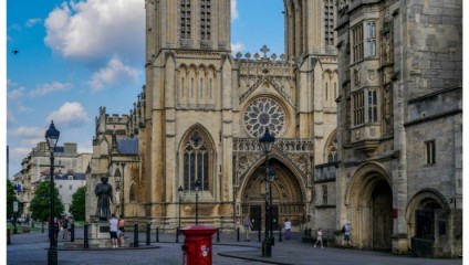 A historic Gothic cathedral with intricate stone carvings and a large rose window is the focal point. People are walking and standing around the plaza in front. A red mailbox and a statue are visible, and the sky is partly cloudy.