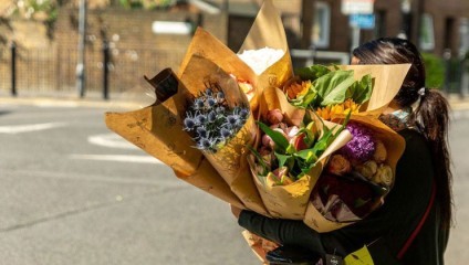 A person with dark hair, seen from behind, carries multiple large bouquets of colorful flowers wrapped in brown paper. The background shows a street scene with buildings and a sunny day.