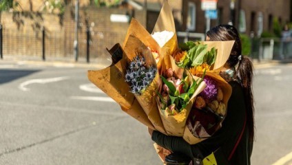 Person with long hair standing outdoors, holding several large bouquets of colorful flowers wrapped in brown paper. Sunlight illuminates the scene, and a street and buildings are visible in the background.