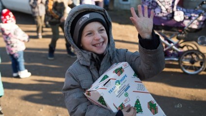 A child in a grey winter coat and hat is smiling brightly, holding a decorated box with TEAM HOPE visible. The child is waving with their other hand. In the background, other children and people are seen, with one pushing a stroller.