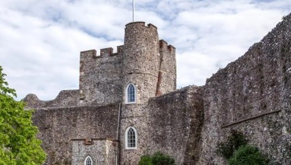 A stone castle with crenellated towers and arched windows stands against a partly cloudy sky. The walls are weathered and covered with some vegetation. There is greenery, including a tree, in the foreground on the left side of the image.