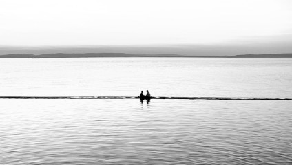 A black and white photo of two people sitting on a narrow beam or ledge extending into a calm body of water. The vast, serene water surrounds them, and distant land is visible on the horizon under a soft, overcast sky.
