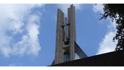 A tall, modern church spire with a cross in the center rises against a partly cloudy blue sky. The structure appears to be made of concrete and has a sleek design with vertical elements. Some tree branches are visible to the right.