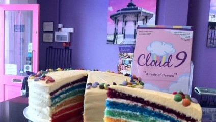 A vibrant bakery scene with a multi-layered rainbow cake topped with white icing and confetti-like decorations. The background features purple walls, a pink door, framed pictures, and a sign reading Cloud 9: A Taste of Heaven with a pastel-colored cloud logo.