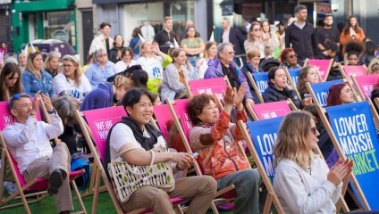 A diverse group of people sitting on colorful deck chairs with banners reading Lower Marsh Market in an outdoor event. Some participants are clapping, smiling, and enjoying the atmosphere. The background shows shopfronts and more people standing or walking around.