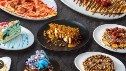 An assortment of colorful desserts displayed on plates, including layered cakes, crepes, pancakes, and pastries, with various toppings like chocolate drizzle, fruits, whipped cream, and sprinkles, arranged on a dark textured surface.