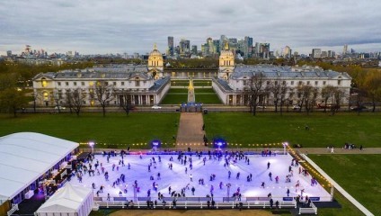 Aerial view of an outdoor ice skating rink filled with skaters. The rink is illuminated by lights, and a large building complex is visible in the background. The sky is overcast, and a cityscape with modern skyscrapers can be seen in the distance.