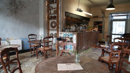 Cozy café interior featuring wooden tables and chairs, rustic wooden countertops, shelves with decorative items, and hanging lights. The space is softly lit, with vases of flowers on tables and a menu displayed on one of the tables in the foreground.