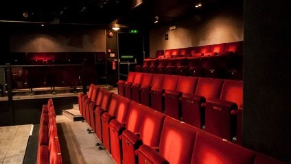 A small theater with rows of red upholstered seats arranged in a stepped formation. The theater is dimly lit, creating a cozy atmosphere. The stage area is visible on the left side, with black walls and ceiling adding to the intimate setting.