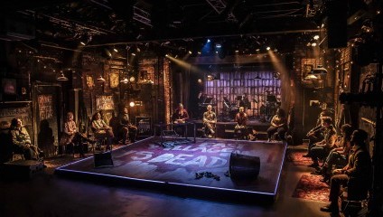 A stage with actors and actresses of RENT sitting around the edge, theatre lights are on