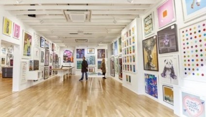 A spacious art gallery with white walls and wooden floors displays a variety of colorful artworks. Two people stand in the gallery, admiring the art pieces hung on the walls. The gallery is well-lit with overhead lights, highlighting the vibrant artwork.