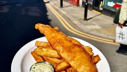 fish and chips with a slice of lemon on a white plate being held up in front of the camera on a road outside Trof