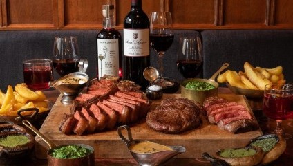 Lots of steak dishes and bottles of red wine on a cluttered table