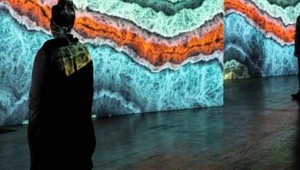 Two people stand in front of an abstract art display featuring vibrant, wavy patterns in shades of blue, orange, and white. The room is dimly lit, drawing focus to the illuminated artwork on the wall. One person faces the artwork closely, while the other stands further away.